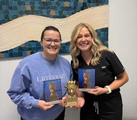 West Madison Dental | Two women smiling and holding plaques and a trophy with a GOAT design inside a dental office.